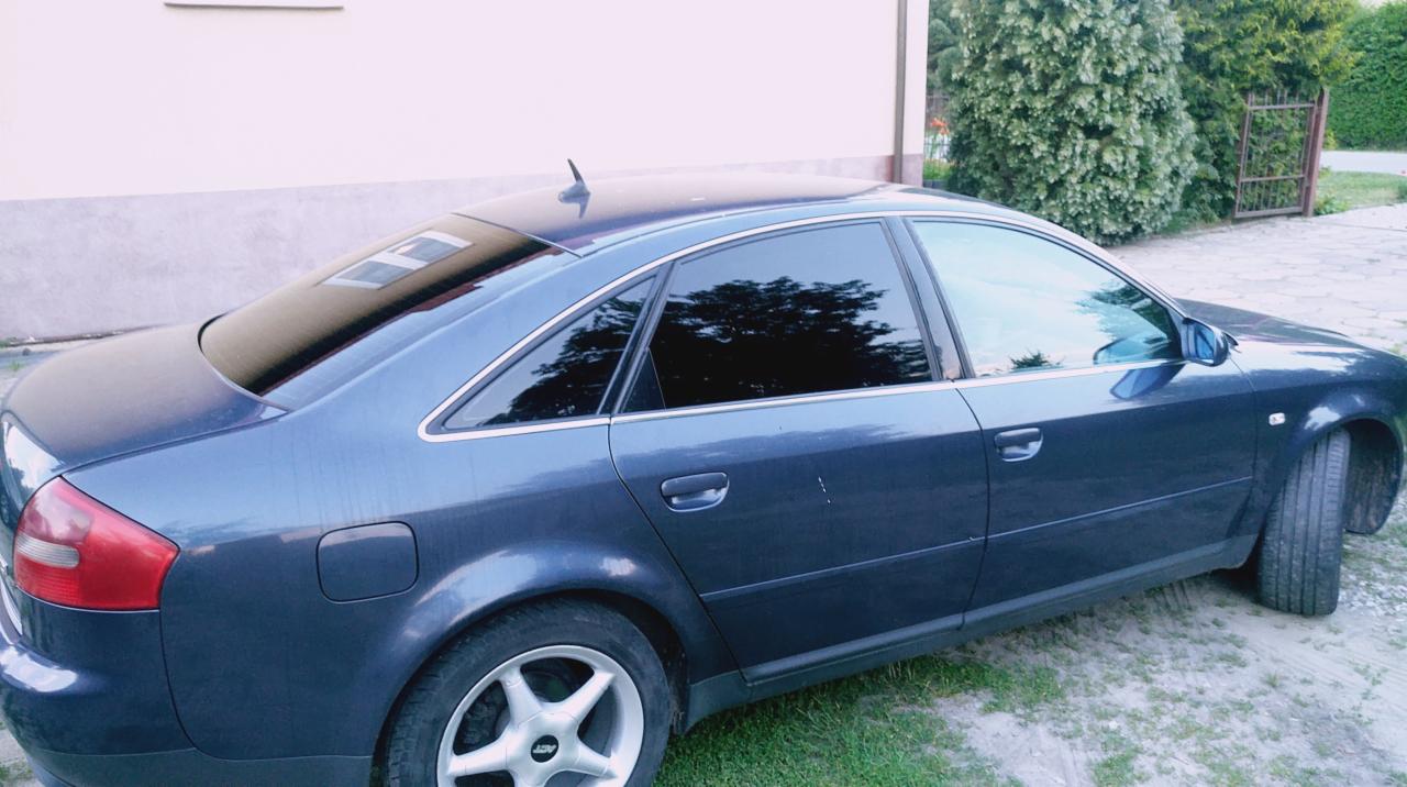 Granatowy sedan Audi A6 z przyciemnionymi szybami bocznymi i tylną, zaparkowany na trawiastym podłożu obok budynku.