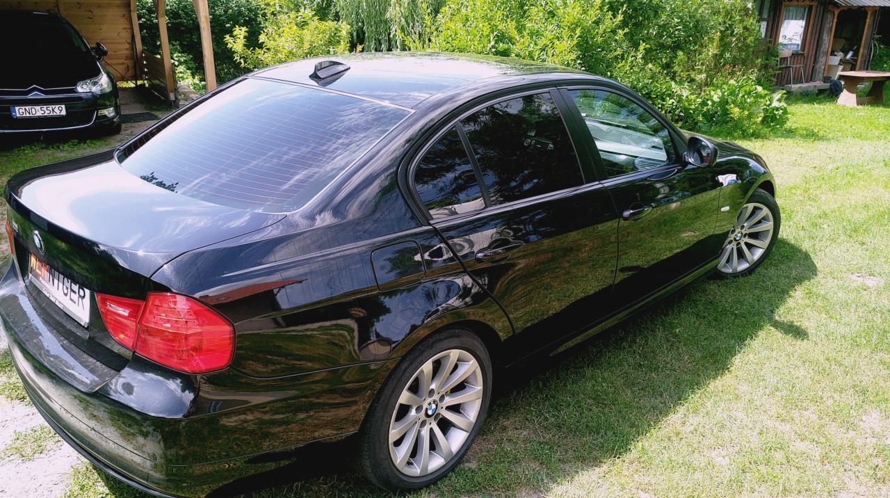 Czarne BMW serii 3 (E90) z przyciemnionymi szybami, zaparkowane na trawniku przed drewnianym domkiem letniskowym, widoczne od strony tylnego błotnika.