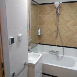 Nowoczesna łazienka z beżowymi płytkami ułożonymi w jodełkę, białą wanną i umywalką z szafką, widoczne chromowane baterie i akcesoria.