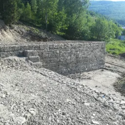Masywna, kamienna ściana oporowa z gabionów na zboczu, zabezpieczająca przed osuwaniem się ziemi, z widocznym krajobrazem górskim w tle.