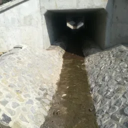 Betonowy przepust drogowy z kamiennym umocnieniem brzegów rowu, widziany z perspektywy wlotu, z widocznym dnem rowu pokrytym wodą i błotem.