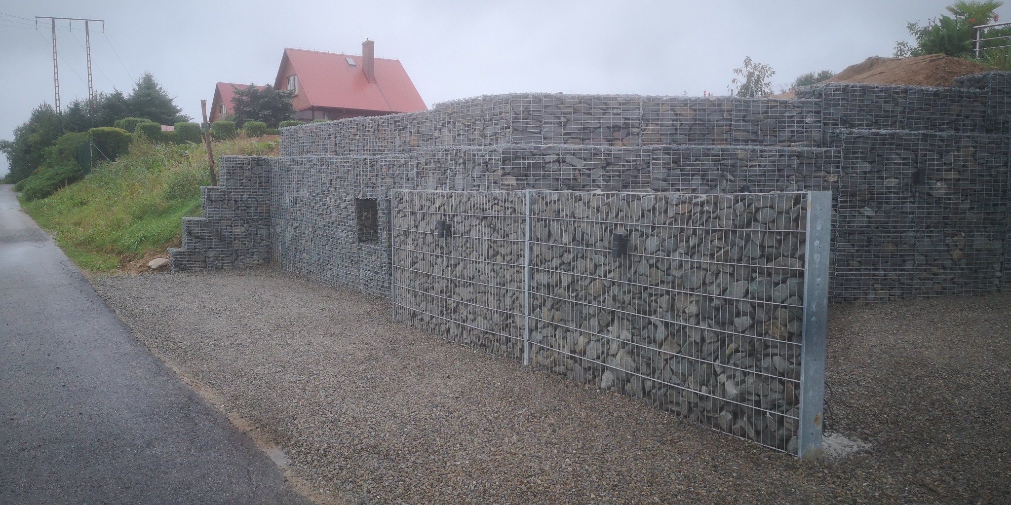 Trójstopniowy mur oporowy z gabionów wypełnionych szarym kamieniem, wzmocniony metalowymi słupkami, otoczony żwirowym podłożem, z widocznymi domami z czerwonymi dachami w tle.