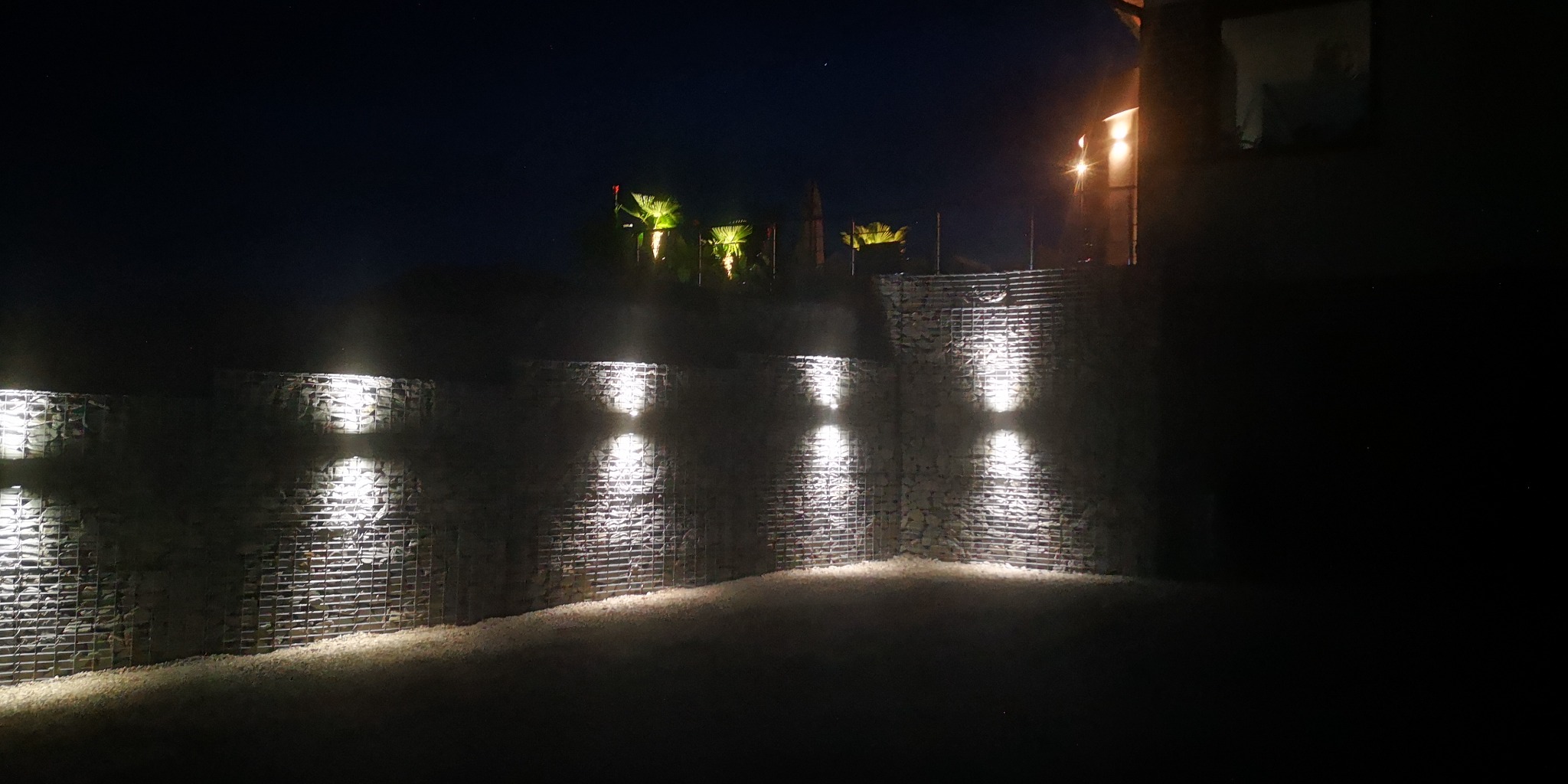 Gabionowa konstrukcja oświetlona punktowymi lampami w nocy, widoczne palmy i budynek w tle.