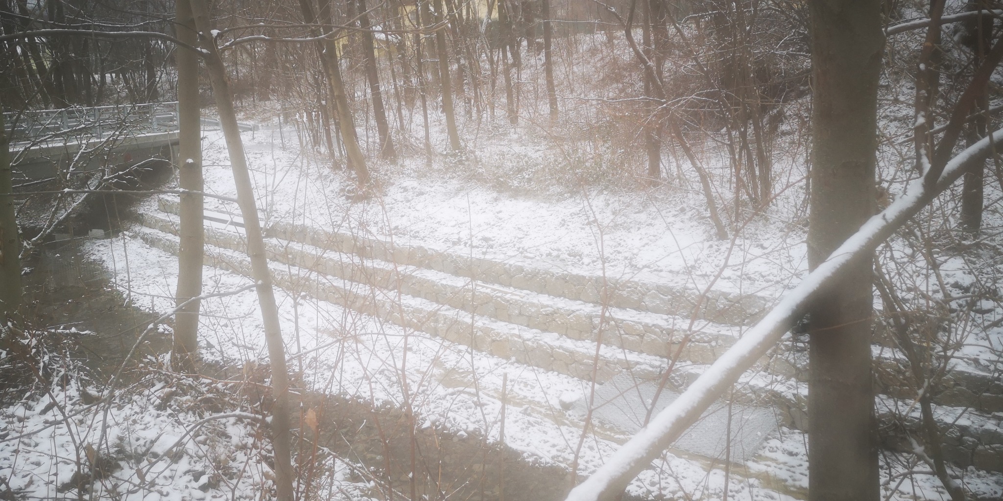 Zimowy krajobraz z potokiem i kaskadowymi umocnieniami brzegów wykonanymi z kamienia, widocznymi spod śniegu, z mostkiem w tle i drzewami na pierwszym planie.