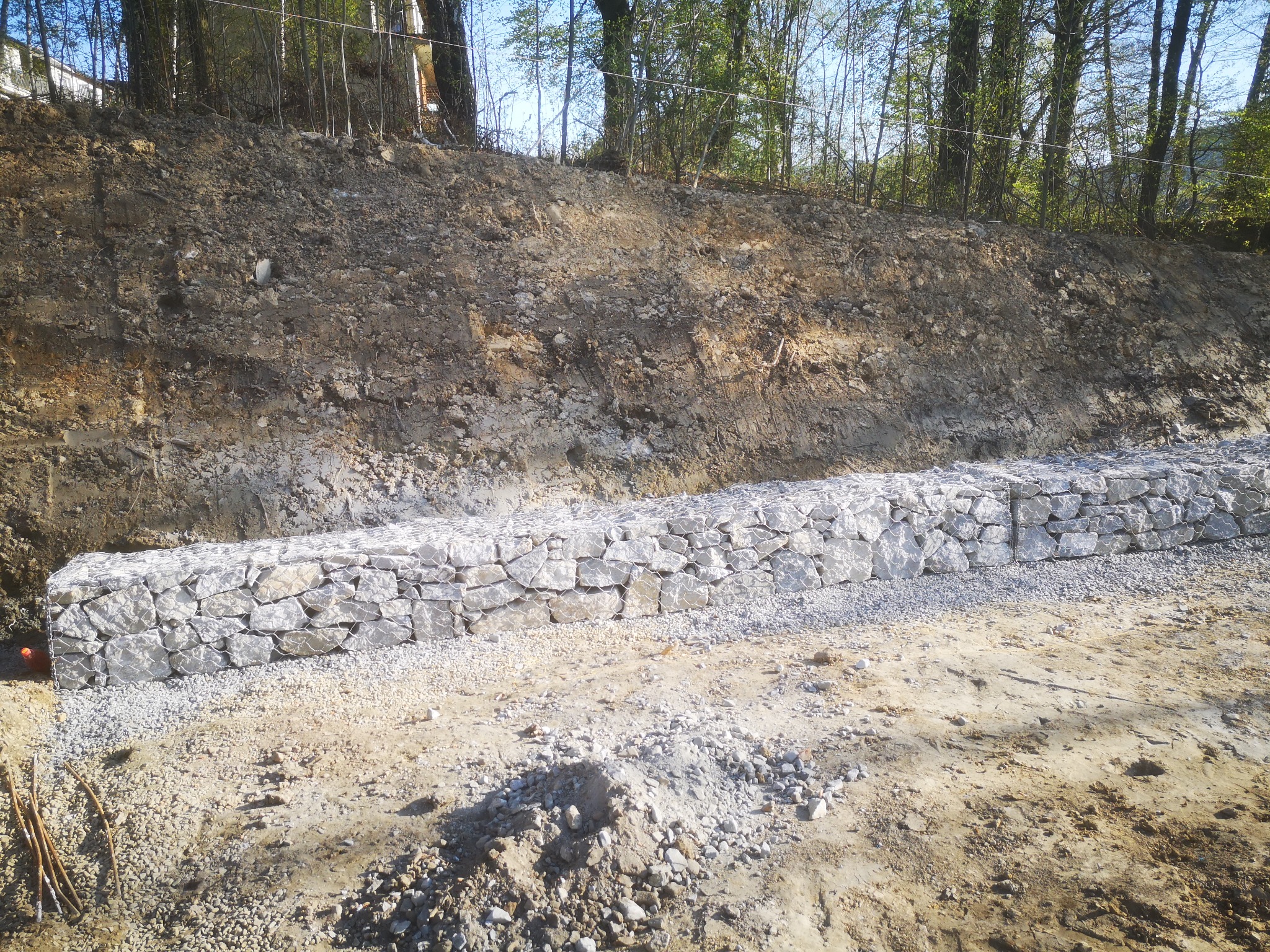 Gabionowy mur oporowy z jasnych kamieni, zabezpieczający skarpę ziemną na tle drzew w słoneczny dzień.