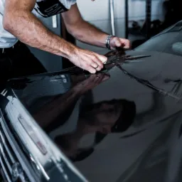 Nakładanie folii ochronnej na maskę czarnego samochodu, widoczne krople wody i odbicie twarzy pracownika.