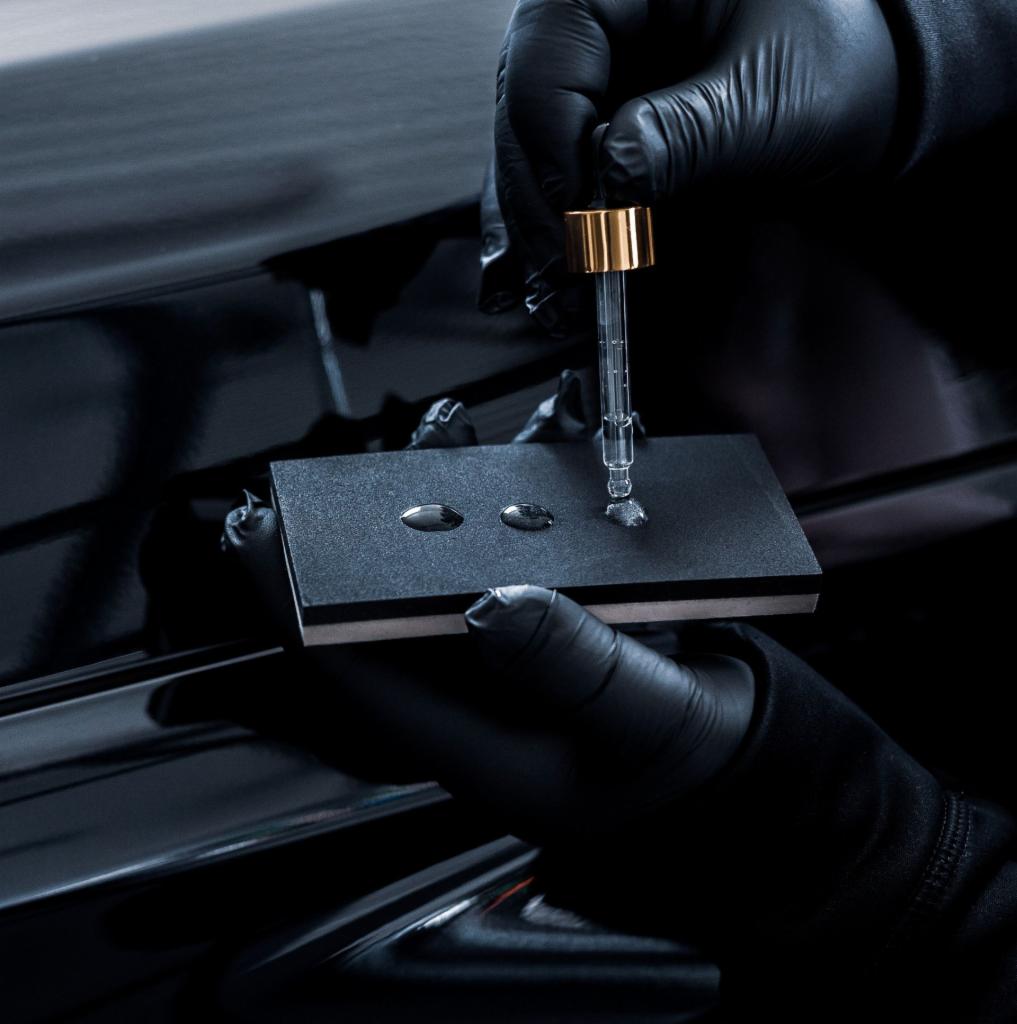 Aplikacja preparatu ochronnego na lakier samochodu przy użyciu pipety i aplikatora, widoczne krople preparatu na czarnym aplikatorze trzymanym w dłoniach w czarnych rękawiczkach, z połyskującym...