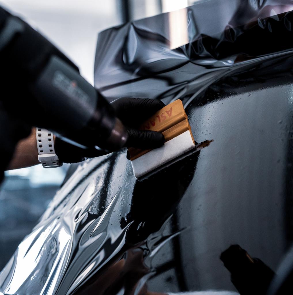 Nakładanie folii ochronnej na karoserię samochodu, widoczna ręka w czarnej rękawiczce wygładzająca folię pomarańczową rakla z logo ASLAN, w tle opalarka.