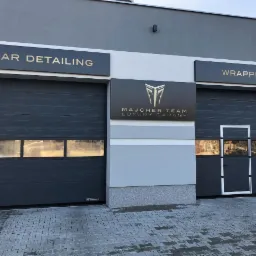 Elewacja garażu: Car Detailing i Wrapping nad szarymi bramami. Logo Majcher Team Luxury Garage. Kostka brukowa na podjeździe. Minimalistyczny design.