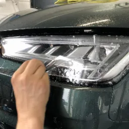 Nakładanie przezroczystej folii ochronnej na reflektor nowoczesnego, ciemnozielonego samochodu, widoczne krople wody na karoserii.