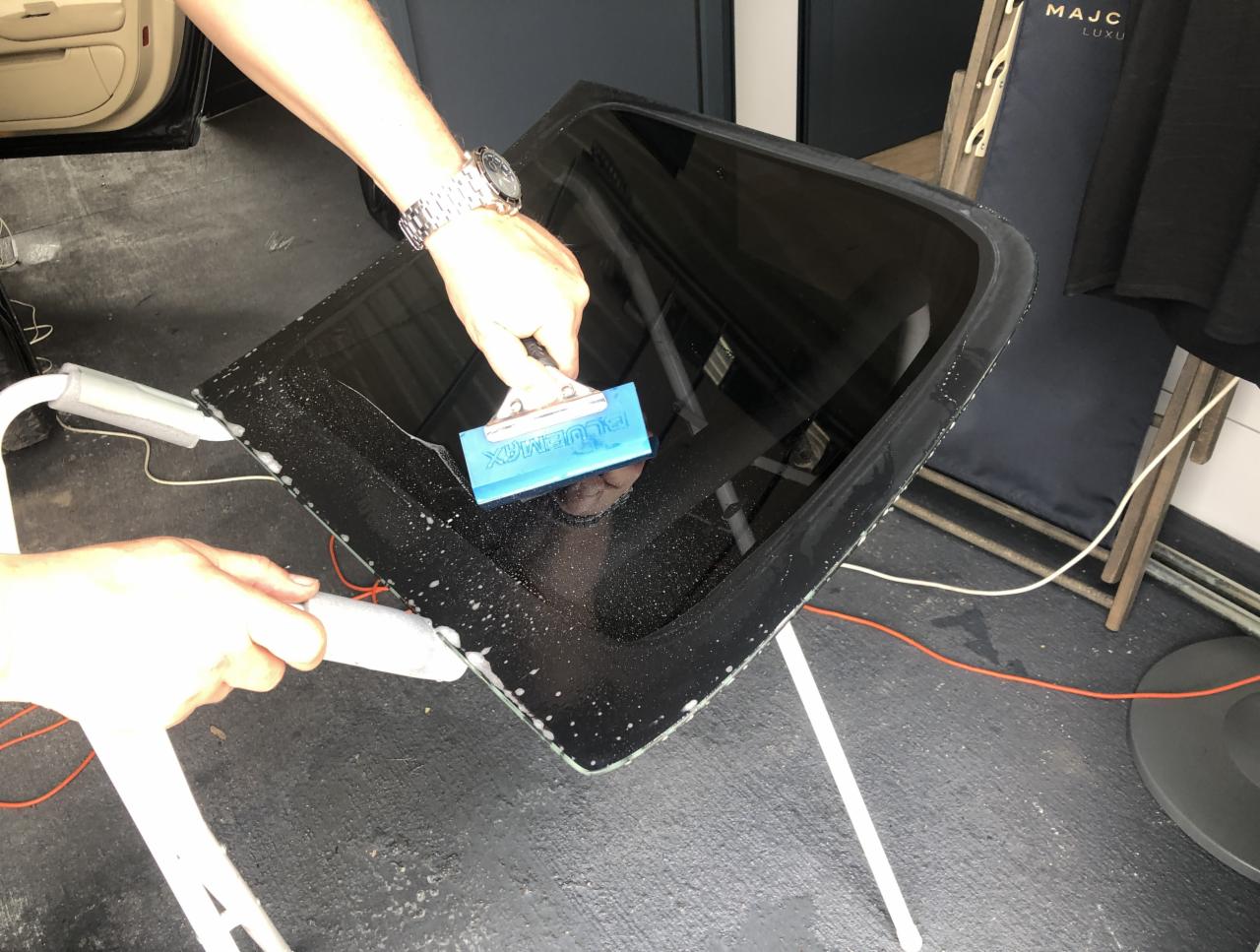 Nakładanie folii na szybę samochodową za pomocą niebieskiej rakli, widoczne drobne pęcherzyki powietrza i woda, w tle fragment wnętrza auta i akcesoria.