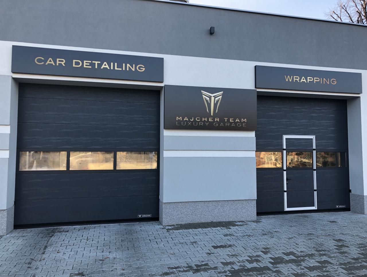Elewacja garażu: Car Detailing i Wrapping nad szarymi bramami. Logo Majcher Team Luxury Garage. Kostka brukowa na podjeździe. Minimalistyczny design.
