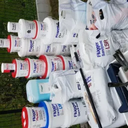 Stos wiader z farbami Flugger różnego typu, worki z gładzią szpachlową Sandplast LSR oraz kuwety malarskie, ułożone na trawie przy ogrodzeniu.