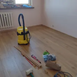 Układanie paneli podłogowych: widoczny fragment ułożonej podłogi, listwy przypodłogowe, poziomica, młotek gumowy, odkurzacz przemysłowy, narzędzia i akcesoria montażowe w pokoju z grzejnikiem.