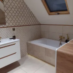 Wykończona łazienka na poddaszu z białą zabudową umywalki, wanną w obudowie z beżowych płytek, złotą armaturą i oknem dachowym.