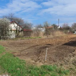 Przygotowany teren pod uprawę, z widocznymi śladami po maszynach rolniczych, w tle dom jednorodzinny i błękitne niebo z chmurami.