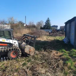 Bobcat z frezem do pni na terenie po wycince, widoczne pozostałości po drzewach, w tle budynek gospodarczy i osoba.