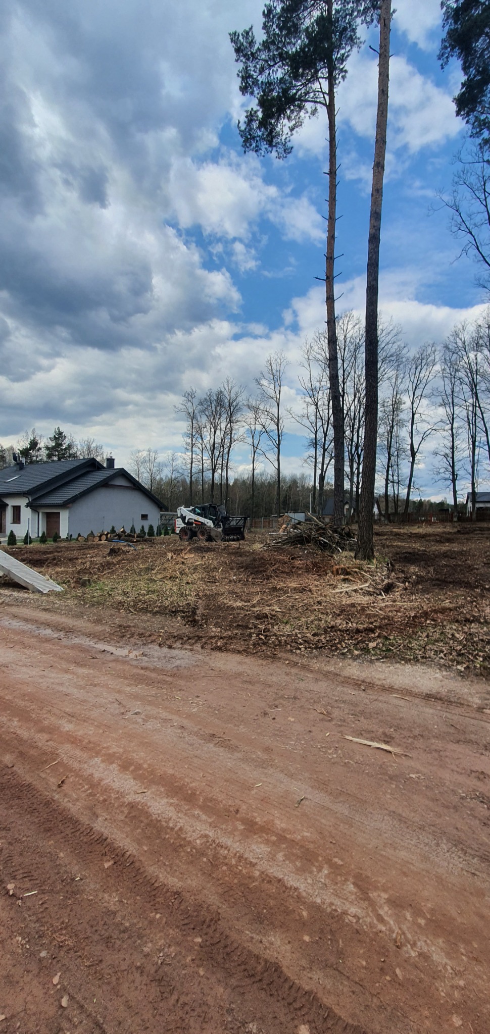 Teren po wycince drzew z widocznym budynkiem mieszkalnym w tle, pozostałości po pniach, gałęzie i maszyna do prac leśnych.