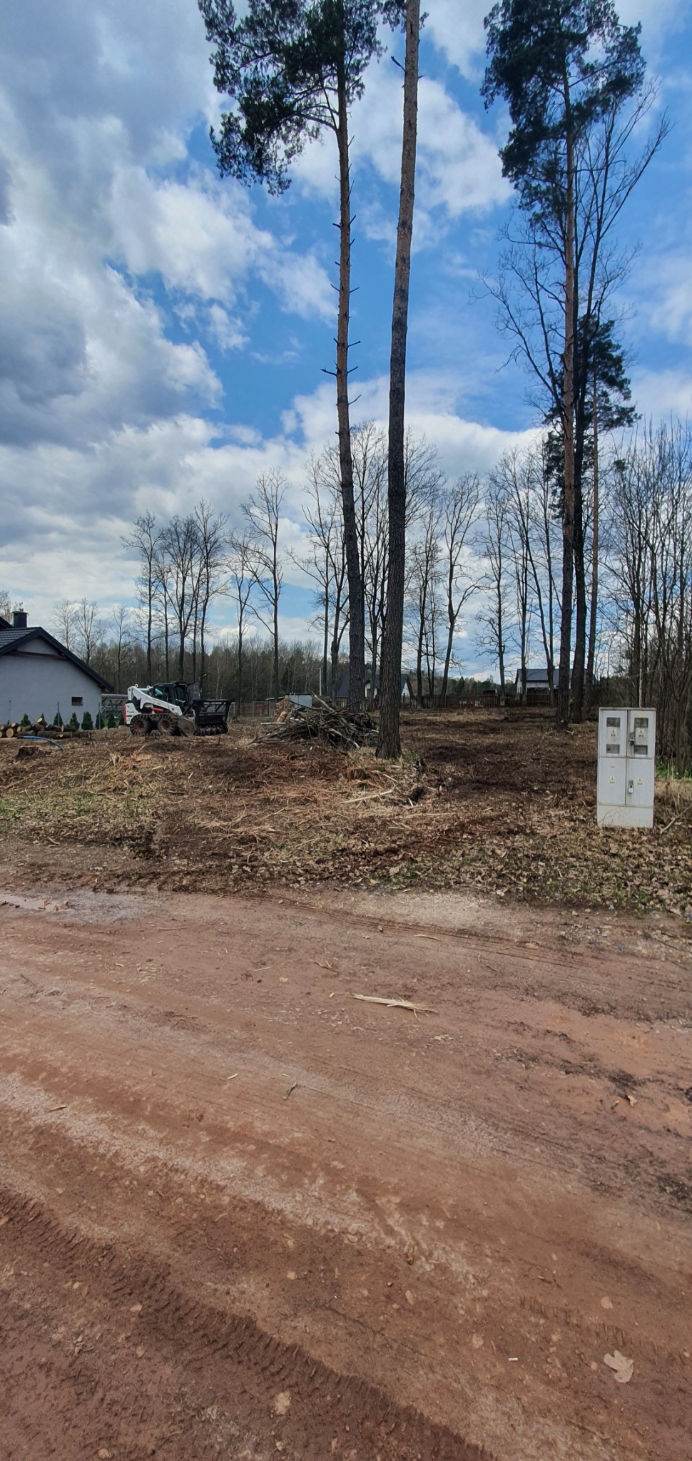 Teren po wycince drzew, z widocznym stosem gałęzi i pni, oraz małą ładowarką Bobcat na tle częściowo zadrzewionego obszaru i budynku mieszkalnego.