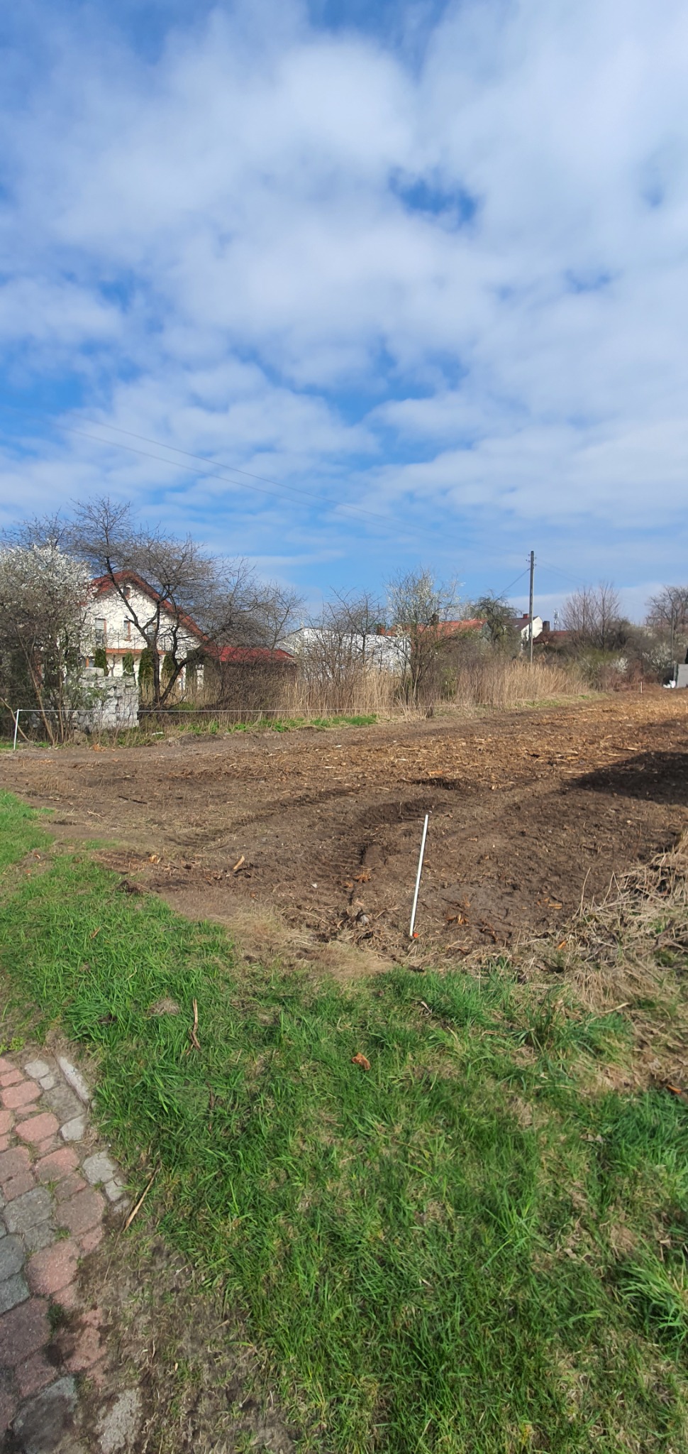Przygotowany teren pod uprawę, z widocznymi śladami po maszynach rolniczych, w tle dom jednorodzinny i błękitne niebo z chmurami.