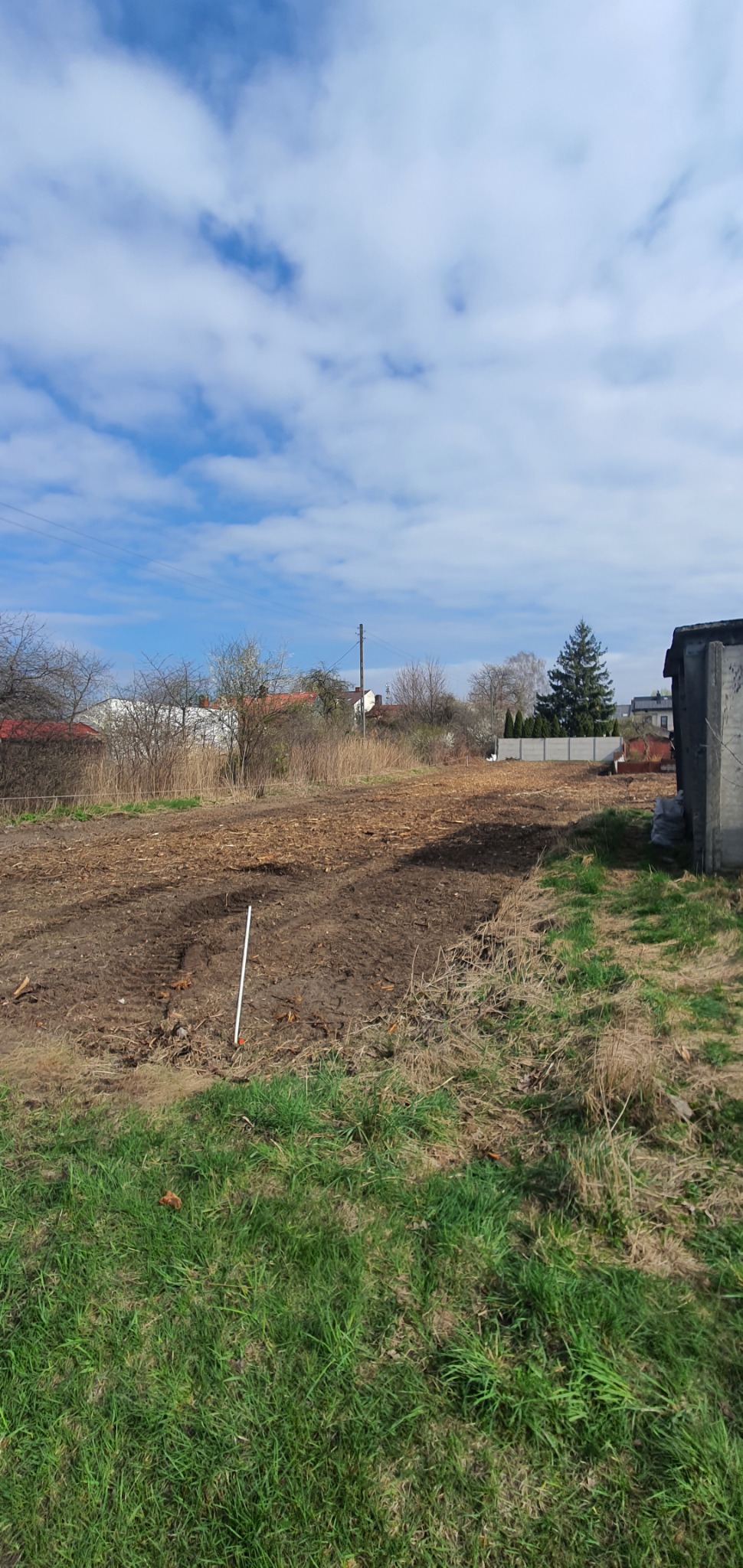 Przygotowany teren pod budowę z widocznymi śladami po maszynach, biały palik wbity w ziemię, w tle zabudowa jednorodzinna i błękitne niebo z chmurami.