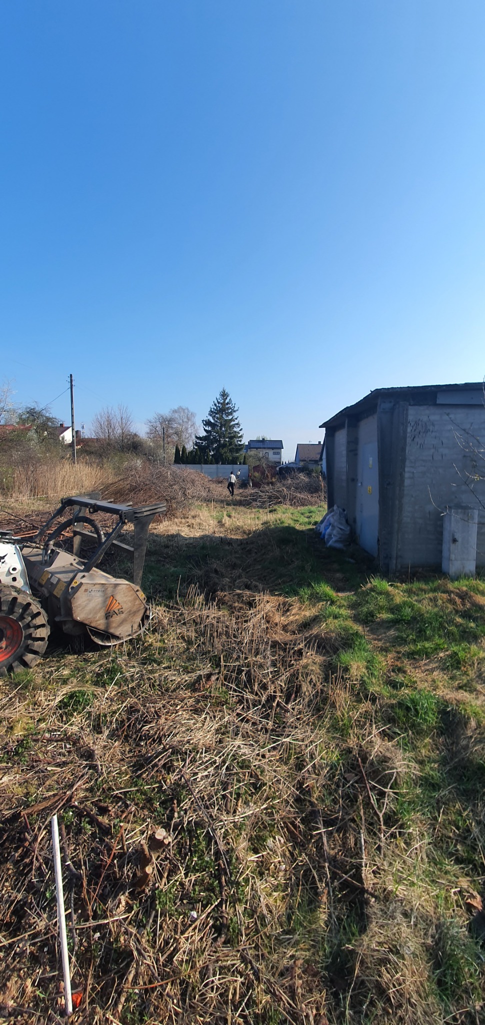 Profesjonalna frezarka do pni w trakcie pracy na zarośniętej działce, z widocznymi resztkami po wycince, w tle budynek gospodarczy i osoba nadzorująca.