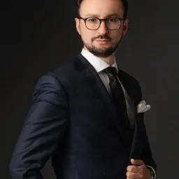 Portret mężczyzny w okularach, w granatowym garniturze z białą poszetką, na ciemnoszarym tle.