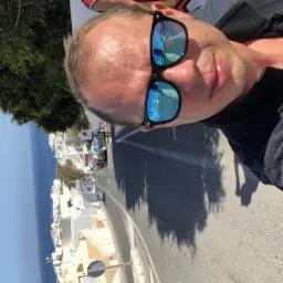 Mężczyzna w okularach przeciwsłonecznych z odbiciem transportu na tle malowniczego krajobrazu miejskiego z morzem w oddali. Fotografia z nietypowej perspektywy.