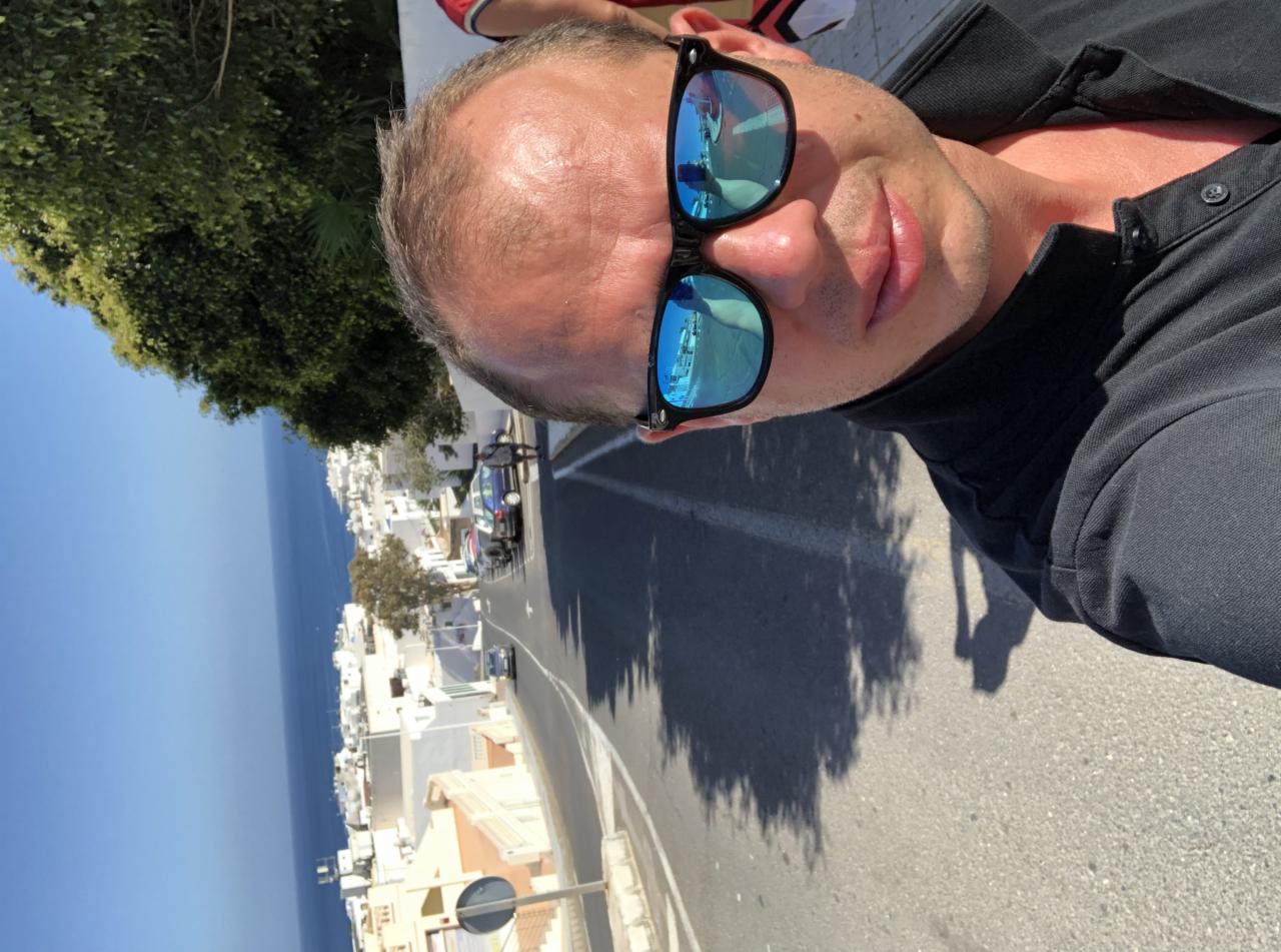 Mężczyzna w okularach przeciwsłonecznych z odbiciem transportu na tle malowniczego krajobrazu miejskiego z morzem w oddali. Fotografia z nietypowej perspektywy.