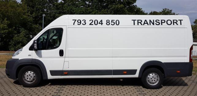 Biały bus dostawczy z napisem 'TRANSPORT' i numerem telefonu 793 204 850, zaparkowany na kostce brukowej, z drzewami w tle.