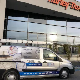 Srebrny samochód dostawczy z drabiną opartą o przeszkloną fasadę budynku Harley-Davidson, z reklamą usług mycia okien, prania wykładzin i czyszczenia posadzek na karoserii.