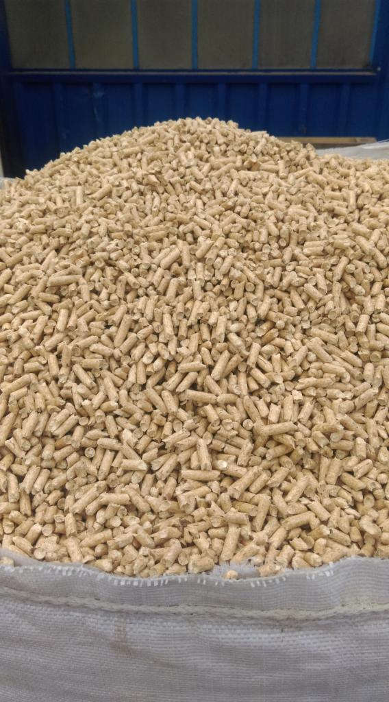 Góra jasnych, cylindrycznych brykietów z biomasy w worku, widoczny fragment niebieskiej konstrukcji w tle.