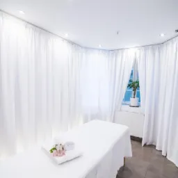 Jasne wnętrze gabinetu z białymi zasłonami, leżanką przykrytą białym prześcieradłem, ręcznikiem i kwiatami; roślina w doniczce na parapecie.