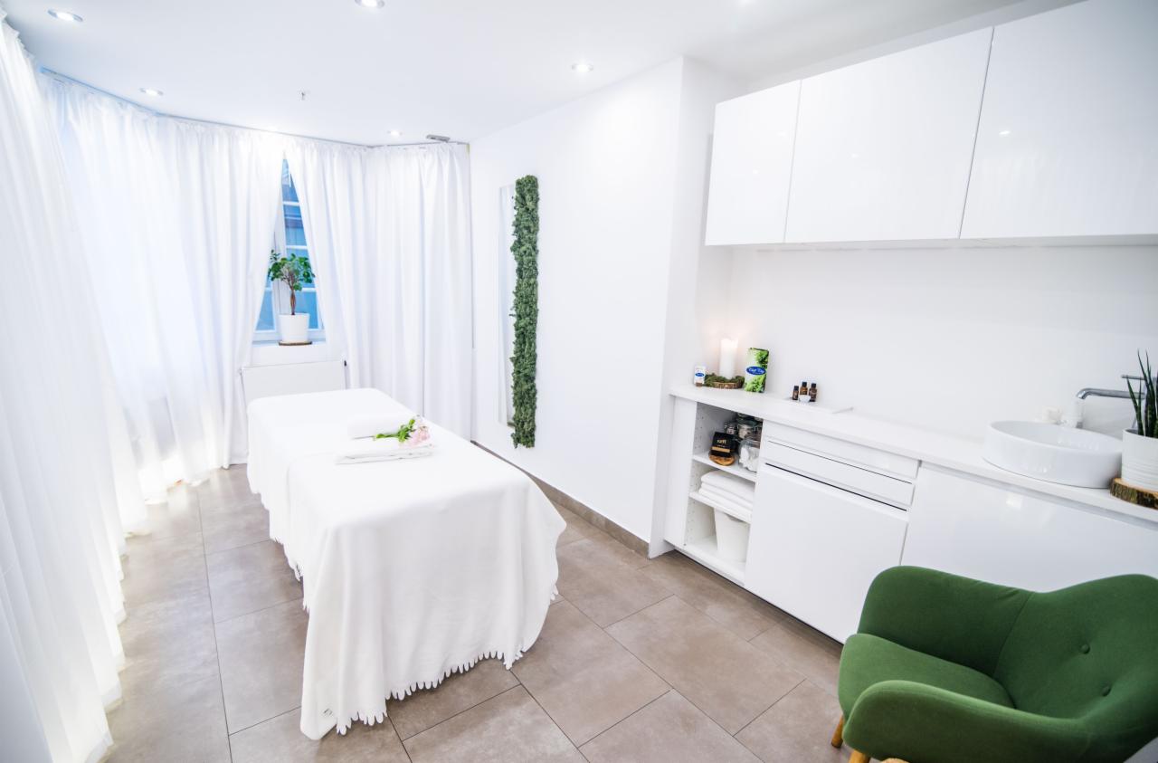 Jasne wnętrze gabinetu z łóżkiem do zabiegów przykrytym białym obrusem z falbanką, na którym leżą zwinięte ręczniki i pojedynczy różowy kwiat. W tle białe szafki, umywalka, zielony fotel...