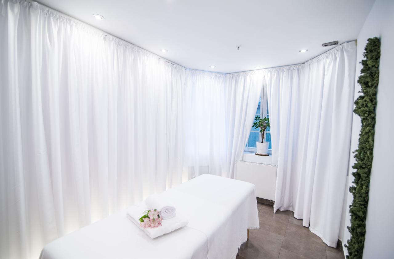 Jasne wnętrze gabinetu z białymi zasłonami, leżanką przykrytą białym prześcieradłem, ręcznikiem i kwiatami; roślina w doniczce na parapecie.