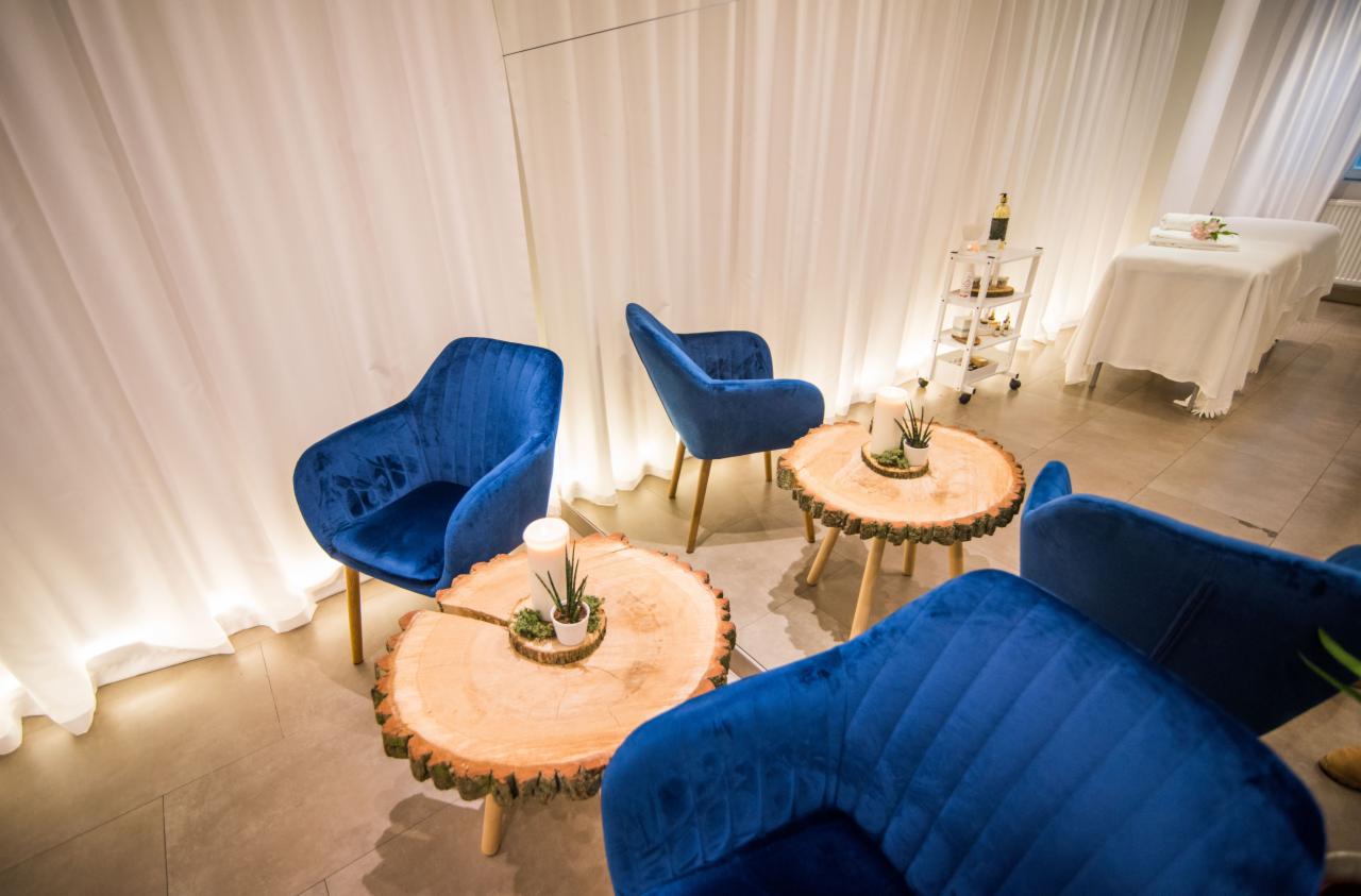 Wnętrze gabinetu z niebieskimi, welurowymi fotelami, stolikami z plastrów drewna, świecami i roślinami w doniczkach, w tle biały parawan i stół do zabiegów z ręcznikami i kwiatami.