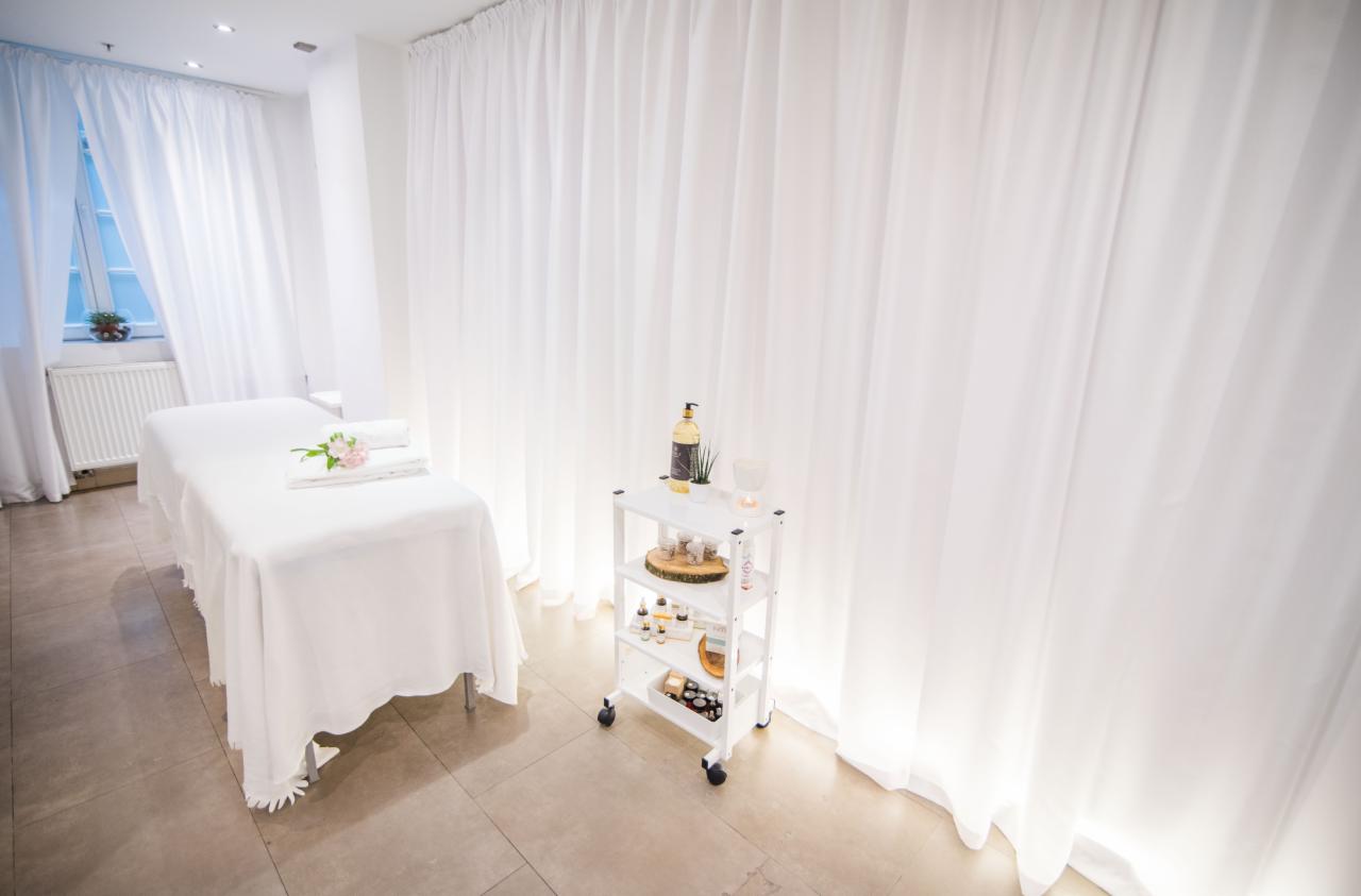 Jasne wnętrze gabinetu z białym łóżkiem do masażu, ręcznikami z różowym kwiatem i wózkiem z olejkami eterycznymi, zasłonięte okna.