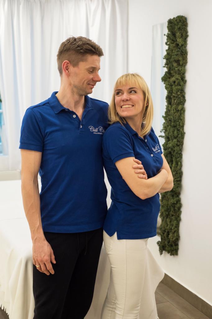 Dwoje pracowników w niebieskich koszulkach polo z logo firmy, stojący obok stołu do masażu w jasnym pomieszczeniu z dekoracją roślinną na ścianie.