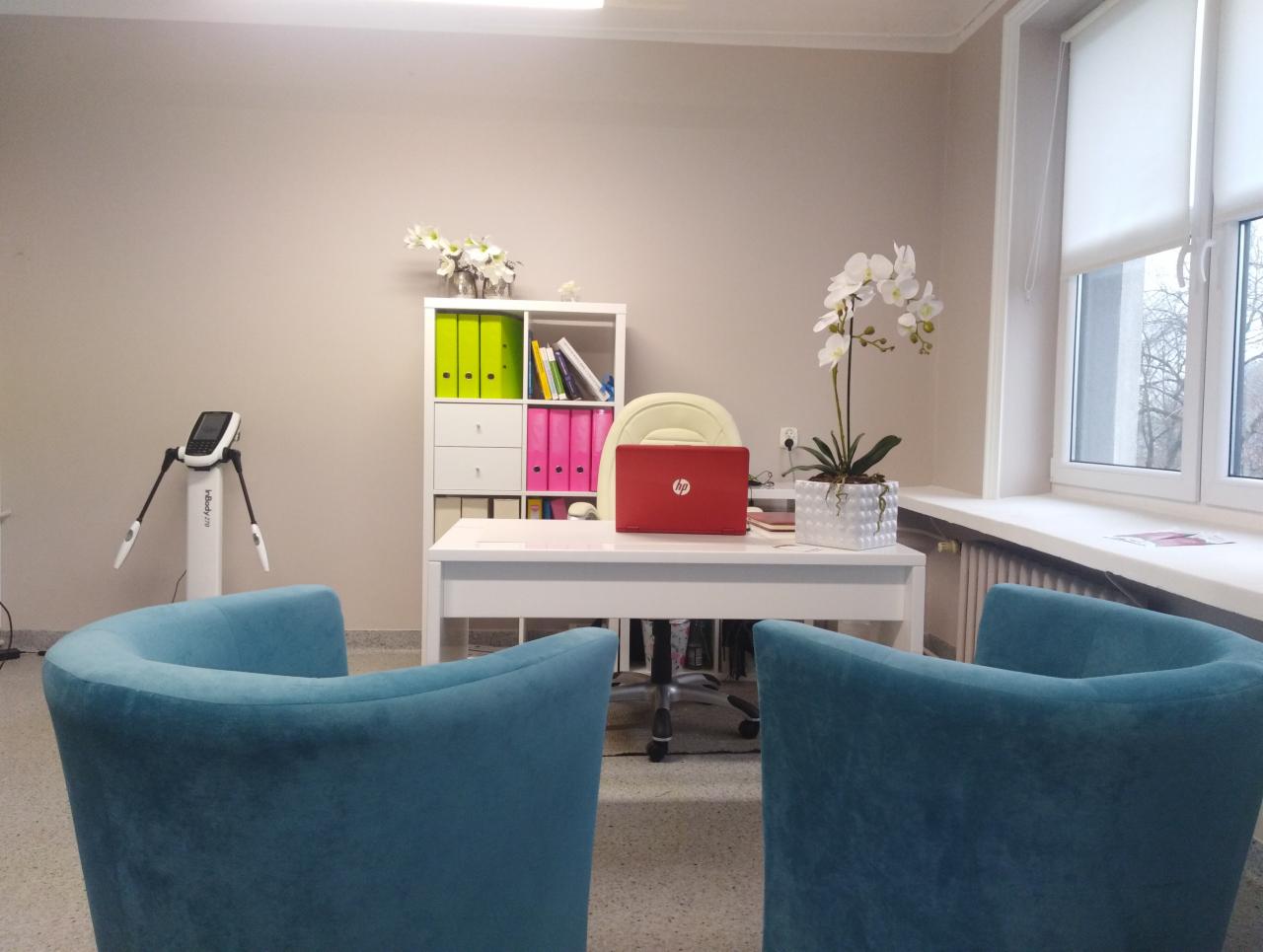 Jasne biuro z białym biurkiem, czerwonym laptopem HP, białą szafką z kolorowymi segregatorami, kwiatami w doniczkach i analizatorem składu ciała InBody 270, z dwoma turkusowymi fotelami...