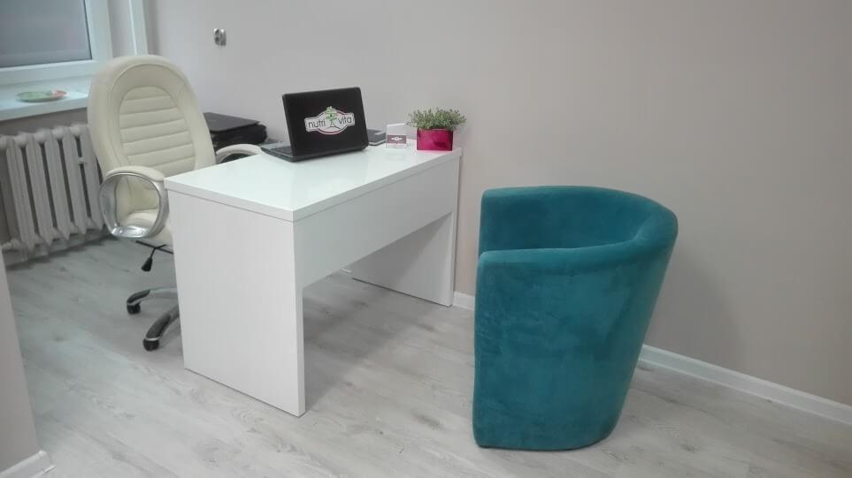 Jasne biuro z białym biurkiem, laptopem z logo Nutri Vita, kremowym krzesłem biurowym i turkusowym fotelem.