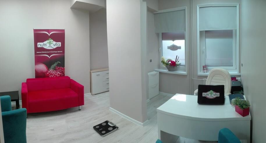 Jasne wnętrze gabinetu z wagą na podłodze, czerwona sofa i roll-up z logo Nutri Vita, biurko z laptopem z tym samym logo.