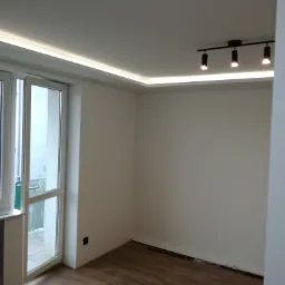 Pokój po remoncie z drewnopodobną podłogą, białymi ścianami, oświetleniem LED na suficie i reflektorami punktowymi, z widocznym wiadrem z farbą i akcesoriami malarskimi na podłodze.