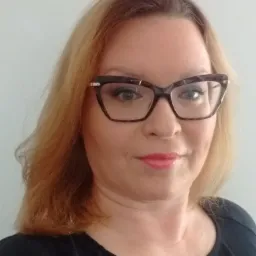 Portret kobiety w okularach o geometrycznych oprawkach, z blond włosami i delikatnym makijażem, na jasnym tle.