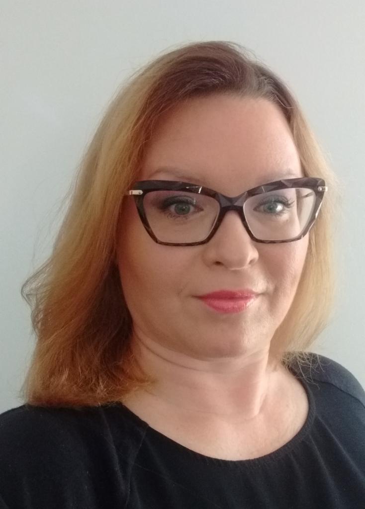 Portret kobiety w okularach o geometrycznych oprawkach, z blond włosami i delikatnym makijażem, na jasnym tle.