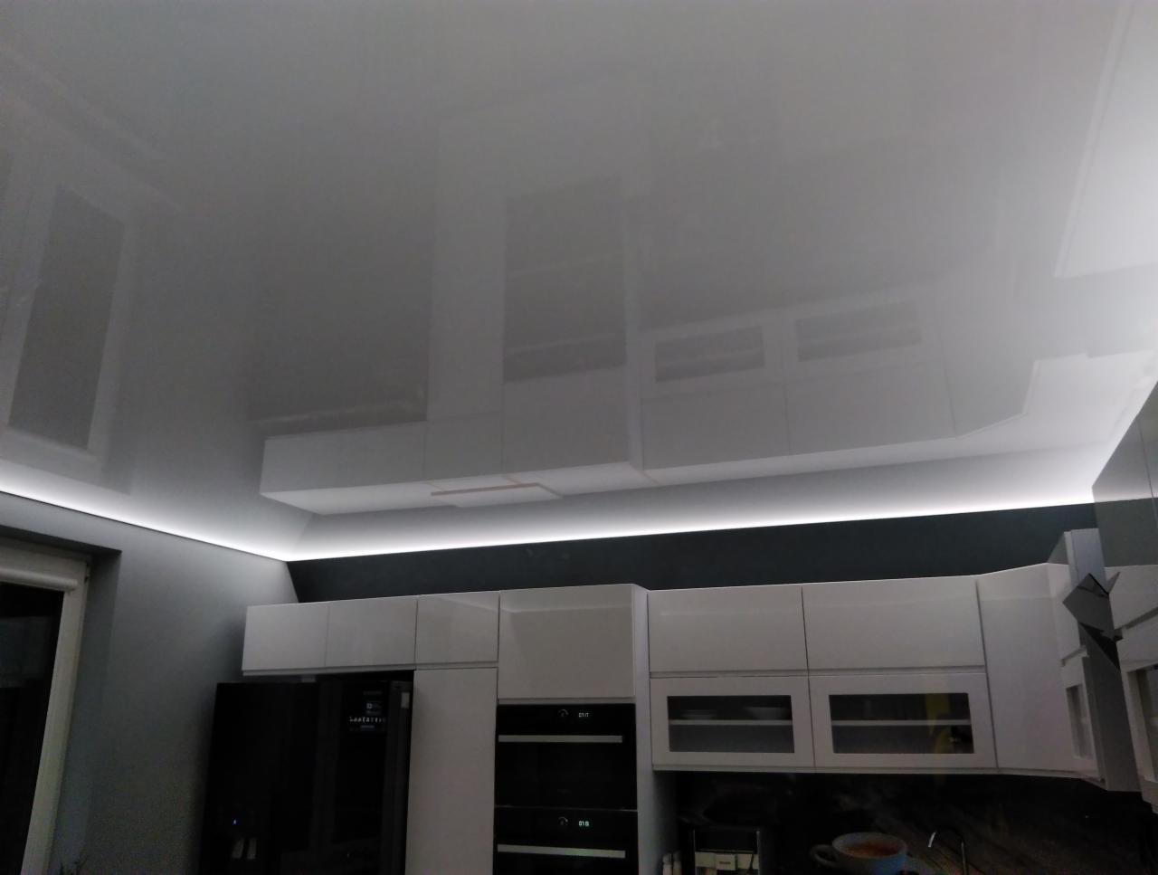 Biały, błyszczący sufit napinany z oświetleniem LED w nowoczesnej kuchni z białymi szafkami i czarnym sprzętem AGD.