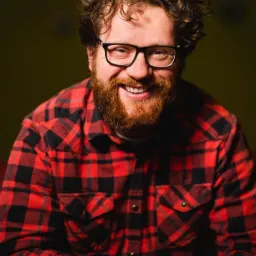 Portret uśmiechniętego mężczyzny z kręconymi włosami i brodą, w okularach i czerwonej koszuli w kratę na ciemnym tle.