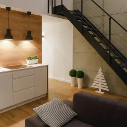 Nowoczesne wnętrze z drewnianą ścianą, białymi szafkami kuchennymi, czarnymi lampami wiszącymi, schodami z metalową konstrukcją i szarym betonem na ścianie, uzupełnione o minimalistyczną dekorację...
