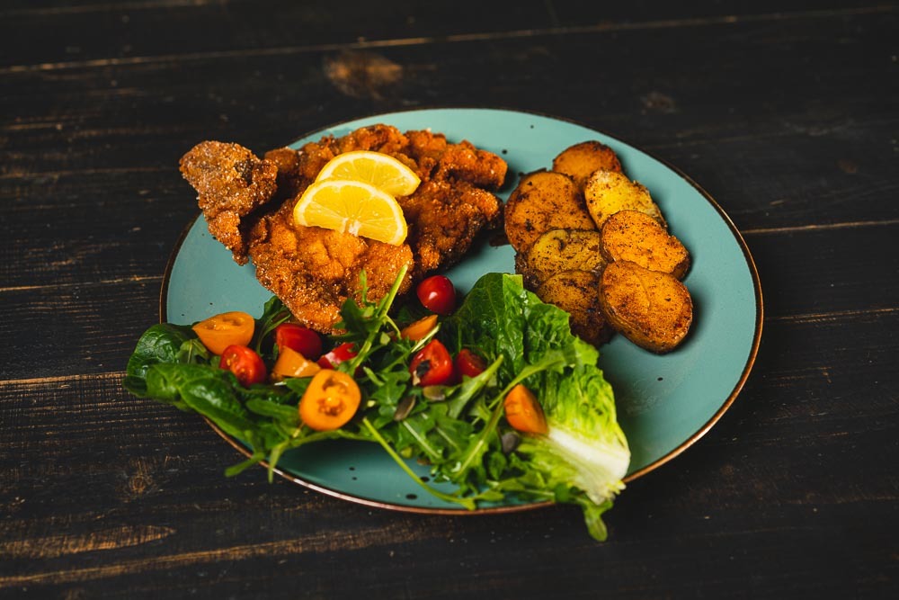Kotlet schabowy z plasterkami cytryny, pieczonymi ziemniakami i sałatką z pomidorkami na turkusowym talerzu z ciemnym, drewnianym tłem.