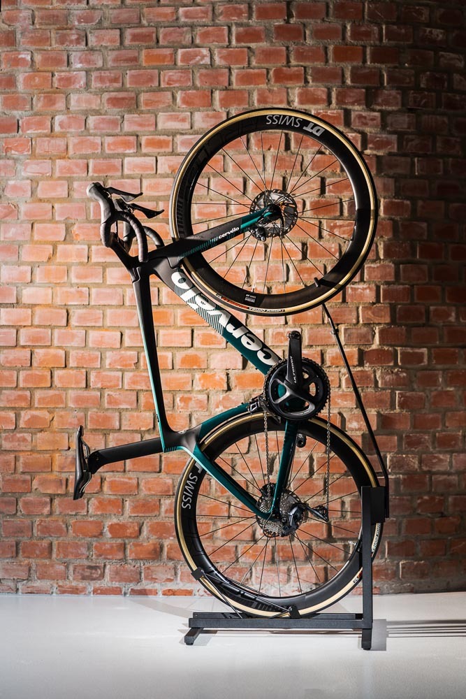 Ciemnozielony rower szosowy Cervelo z karbonową ramą, ustawiony pionowo na stojaku przed ścianą z czerwonej cegły.