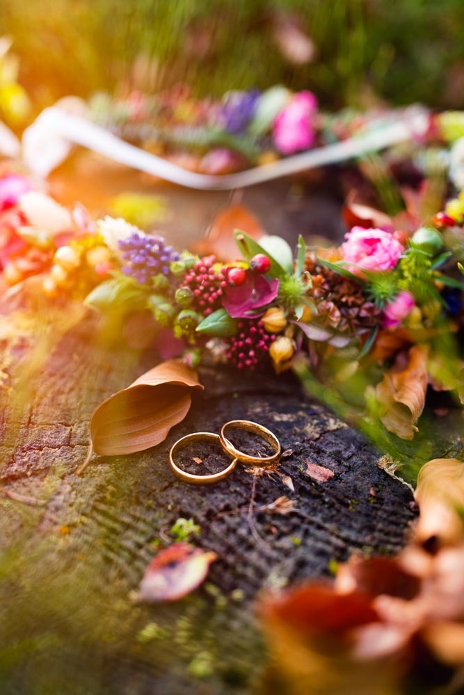 Dwa złote obrączki ślubne leżące na spróchniałym pniu drzewa, otoczone jesiennymi liśćmi i wiankiem z kolorowych kwiatów.